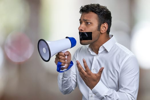 Young man activist with black tape over mouth can not speak into megaphone. Abstract bokeh background.