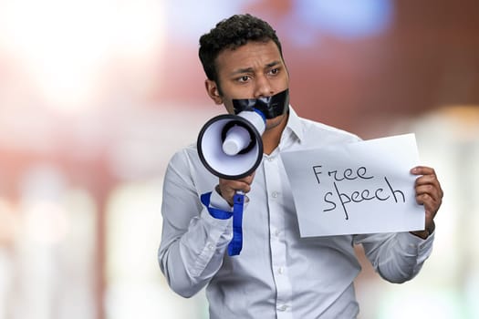 Young indian businessman with sealed mouth holding poster with inscription Free speech. Abstract bokeh background.