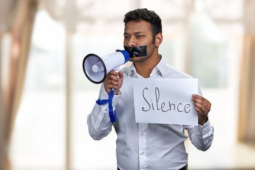 Indian man with taped mouth holding poster with inscription Silence. Censored male protester with megaphone. Freedom of speech.