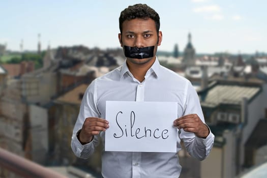 Indian man with taped mouth holding poster with inscription Silence. Blur city buildings in the background.