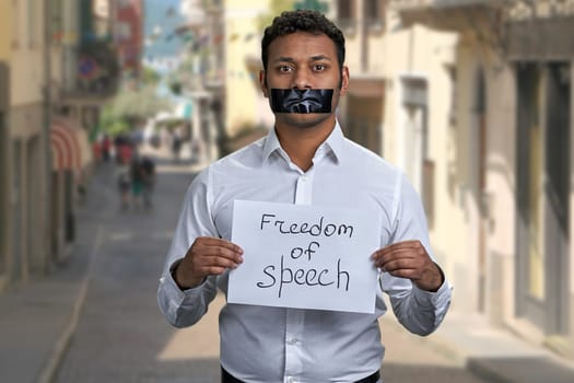 Young man with taped mouth holding paper card with inscription Freedom of speech. Blur narrow city street in the background.