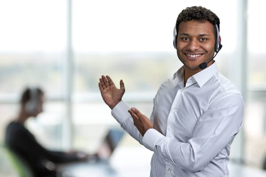 Smiling male call center agent showing something with hands. Blur office interior in the background.