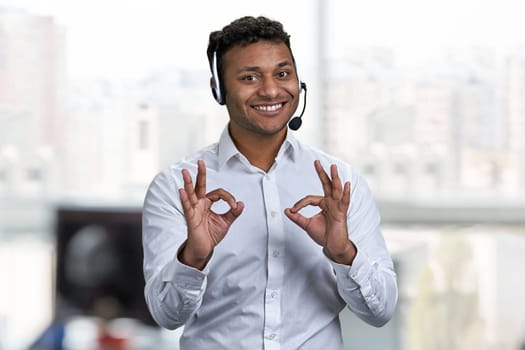 Cheerful male call center operator gesturing okey with both hands. Blur office interior in the background.