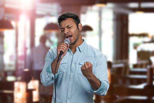 Enthusiastic young indian man performing song with microphone. Interior blur background.