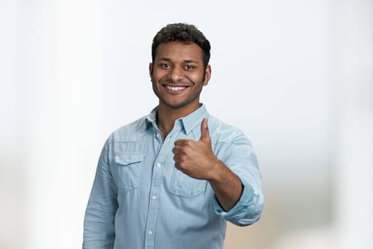 Smiling young man in blue shirt showing thumb up gesture. Attractive guy gesturing thumb up to camera on blurred background.