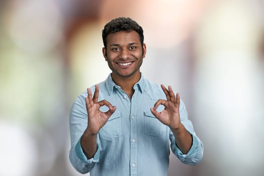 Positive smiling young man showing okey gesture with both hands. Handsome guy gesturing okey on abstract bokeh background.