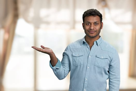 Handsome dark-skinned young man showing empty copy space. Smiling man presenting product on blur interior background.