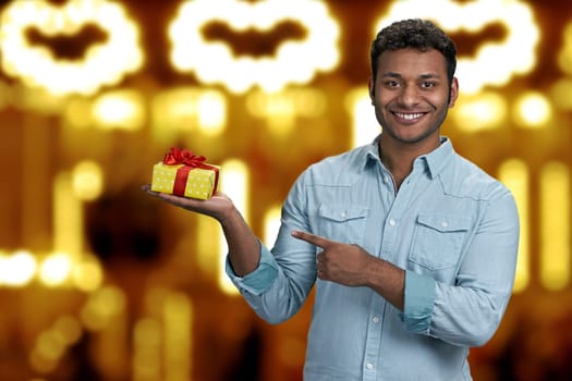 Cheerful young man showing gift box in his hand. Golden bokeh lights background. Christmas holiday sales.