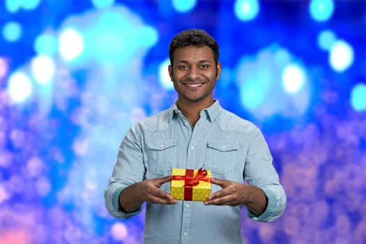 Attractive smiling young man handing gift box to camera. Blue bokeh lights background. Christmas present delivery in time.