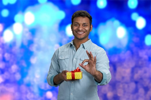 Cheerful young man holding gift box and showing ok gesture. Blue bokeh lights background. Time to celebrate New Year.