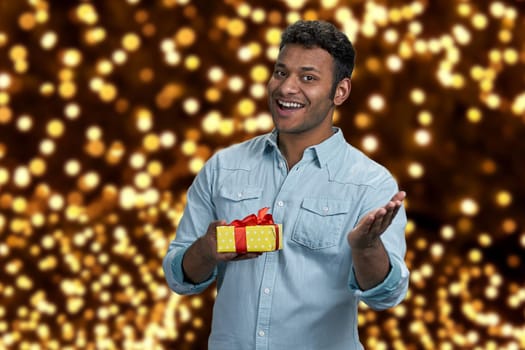 Delighted young man showing gift box and looking at camera. Festive bokeh background. Best Christmas gift shop.