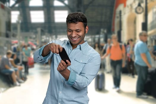 Cheerful young man palying game on his smartphone. Blur background of train station with walking people.