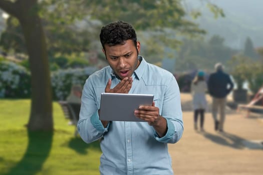 Young shocked man using digital tablet standing outdoors. People, technology and emotions concept.