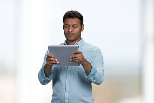Confident young man wearing blue shirt looking at digital tablet. Handsome indian man using computer tablet on blurred background.