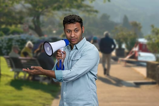 Young indian man holding megaphone and showing something with hand. Blur crowded city street in the background.