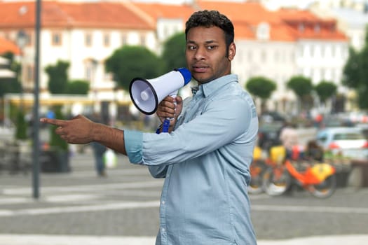 Young indian man talking into megaphone and pointing with index finger. Blur city street in the background.