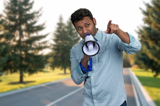 Young indian man talking into megaphone and pointing with index finger. Man with loudspeaker standing outdoors.