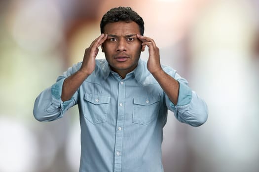 Stressed shocked young man touching his temples because of headache. Abstract bokeh background.