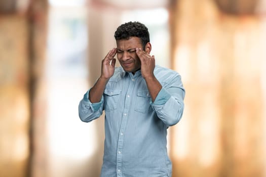 Stressed indian man massaging his temples because of terrible migraine. Exhausted young man standing on blur interior background.