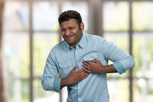 Young indian man suffering from heart ache. Blur interior background. Heart disease concept.