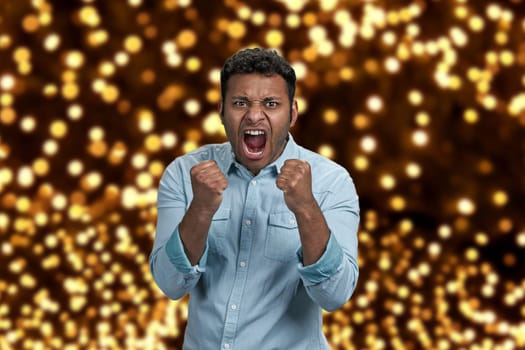 Aggressive young man with clenched fists screaming on festive bokeh lights background. Human negative emotions.