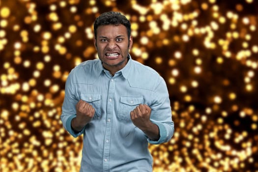 Angry aggressive guy grimacing and shaking fists. Festive bokeh background. Human negative expressions.