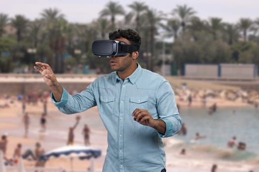 Handsome indian guy wearing virtual reality glasses. Blur swimming pool with tourists and palms in the background.