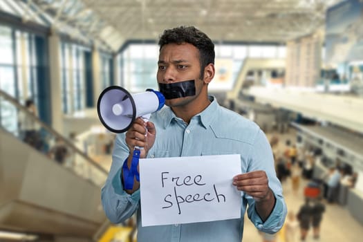 Young man with taped mouth trying to speak into megaphone while standing on escalator at airport. Censored man holding card with inscription Free speech while standing on the sliding walkway in the airport.