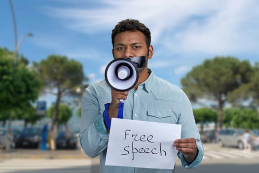 Young man with taped mouth trying to speak into megaphone standing outdoors. Free speech concept.