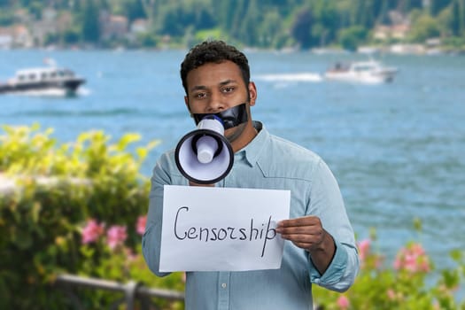 Young man with black tape over mouth and megaphone standing on sea blur background. Censorship concept.