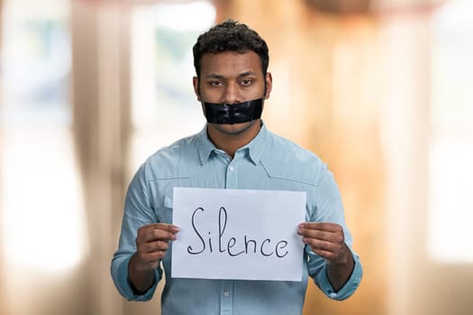 Dark-skinned censored man holding paper banner with inscription Silcence. Interior blur background.