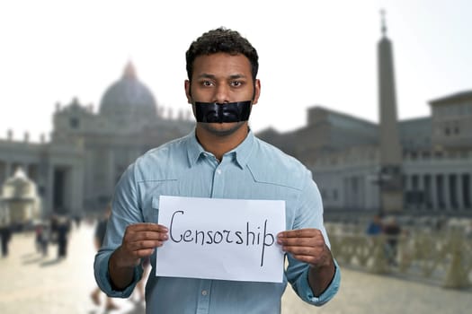 Young indian man with taped mouth holding paper card with handwritten inscription Censorship. City architecture blur background.
