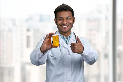 Young smiling indian doctor showing pills and giving thumb up gesture. People, medicine and pharmacy concept.