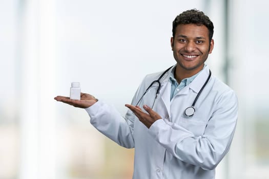 Handsome smiling doctor with stethoscope presenting pills on blur clinic interior background. People, medicine and pharmacology concept.