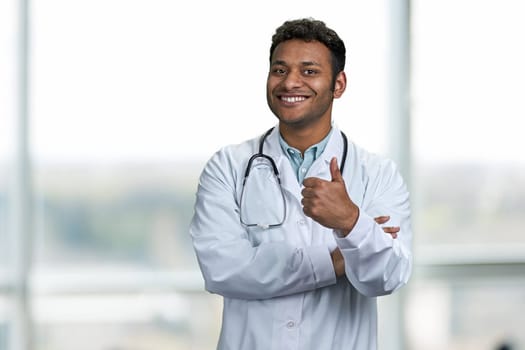 Handsome young doctor with stethoscope giving thumb up to camera. Smiling male doctor looking at camera.