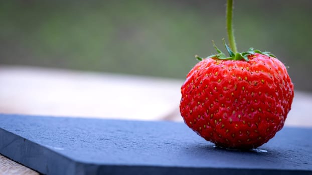 Close up of one strawberry on small black cutting board isolated outdoor on wooden table.