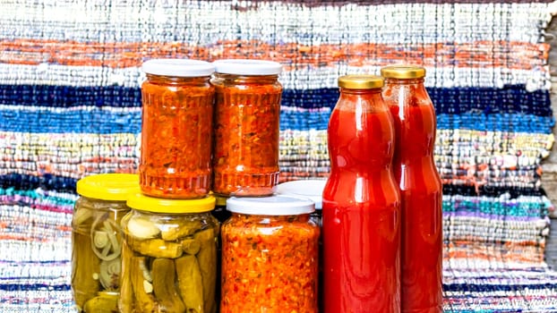 Glass jars with pickled red bell peppers and pickled cucumbers (pickles) isolated. Jars with variety of pickled vegetables, jars with zacusca and bottles with tomatoes sauce. Preserved food concept.