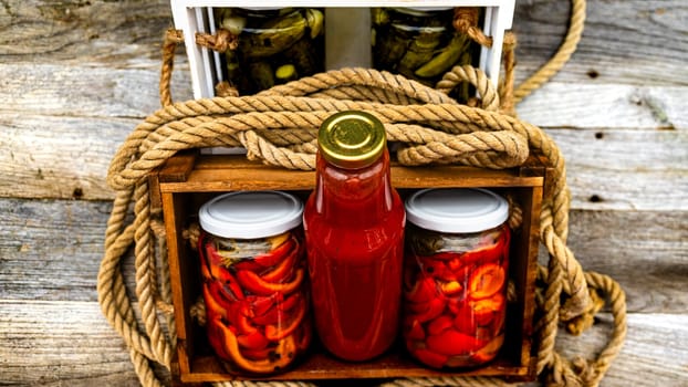 Wooden crate with bottles with tomatoes sauce and glass jars with pickled red bell peppers isolated in a rustic composition. Jars with variety of pickled vegetables preserved food concept.