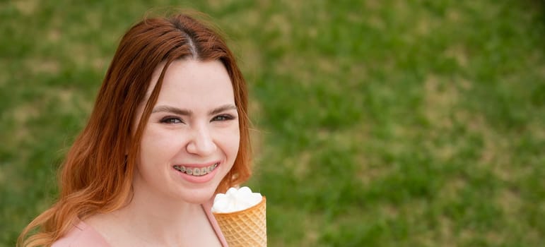 Young beautiful red-haired woman smiling with braces and eating an ice cream cone outdoors. Widescreen