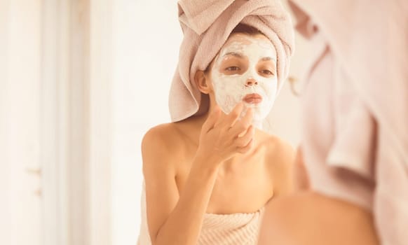 A young girl in a towel on her head and body stands near the mirror in the bathroom and applies a clay mask to her face, the woman takes care of her health and beauty.