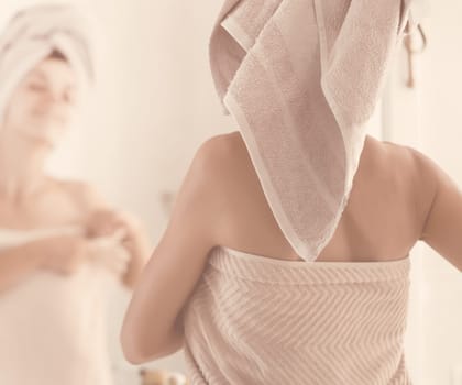 A young cheerful girl in a towel on her head and body stands near the mirror in the bathroom and takes care of her health and beauty.