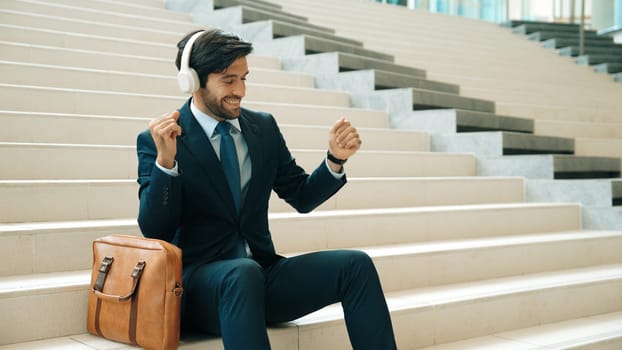 Professional business man picking and listening music by using headphone while sitting at stairs. Project manager dance while listening song while wearing headphone, suit. White background. Exultant.