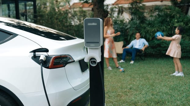 Electric vehicle recharge from home charging station on background of happy and playful family playing together. EV car using alternative and sustainable energy for better future Synchronos