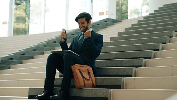 Successful business man celebrate successful project while sitting at stairs. Smart project manager getting new gob, getting promotion, increasing sales while calling friends by using phone. Exultant.