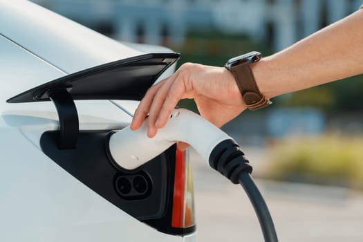 Closeup hand put EV charger to recharge electric car's battery from charging station in city commercial parking lot. Rechargeable EV car for sustainable environmental friendly urban travel. Expedient