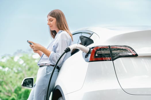 Young woman using smartphone online banking application to pay for electric car battery charging from EV charging station during vacation holiday road trip at national park or summer forest. Exalt