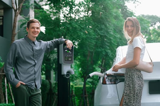 Young couple travel with EV electric car charging in green sustainable city outdoor garden in summer shows urban sustainability lifestyle by green clean rechargeable energy of electric vehicle innards