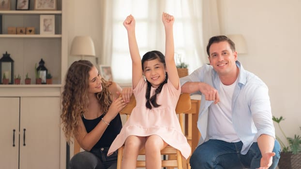 Happy family portrait with lovely little girl smiling and looking at camera, lovely and cheerful parent and their daughter sitting together in living room at home with warm daylight. Synchronos