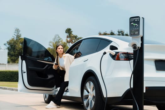 Young woman use smartphone to pay for electricity at public EV car charging station green city park. Modern environmental and sustainable urban lifestyle with EV vehicle. Expedient