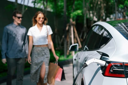 Young couple travel with EV electric car charging in green sustainable city outdoor garden in summer shows urban sustainability lifestyle by green clean rechargeable energy of electric vehicle innards